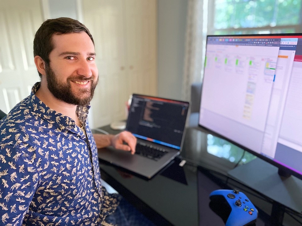 Head shot of Dan in a blue collared shirt with a beta fish pattern on it, sitting at a desk with a MacBook, monitor, and blue Xbox One controller on it
